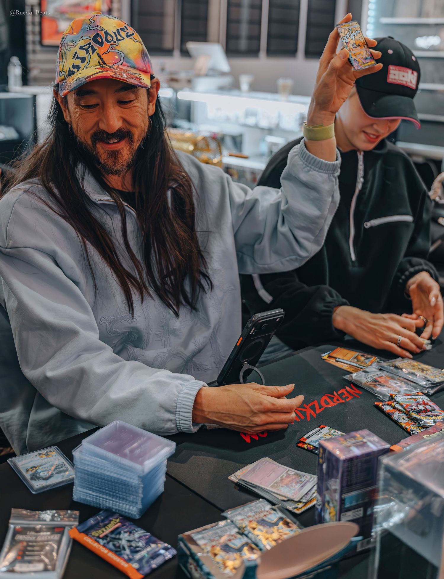 Man in colorful cap opening trading card packs at a collectibles store table.