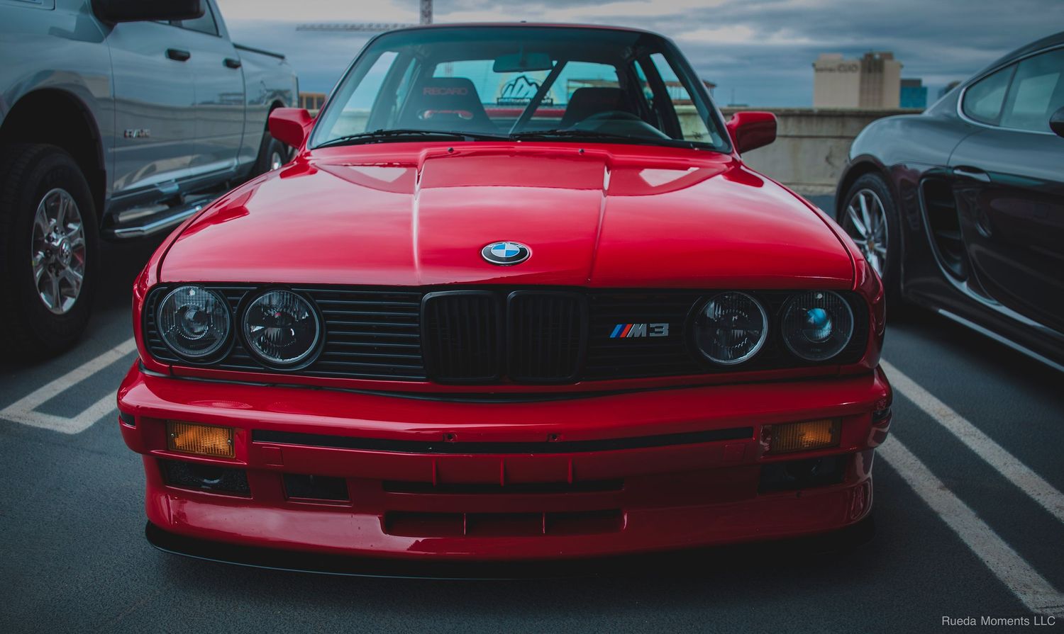 Front view of a red BMW E30 M3 with classic round headlights at a car meet.
