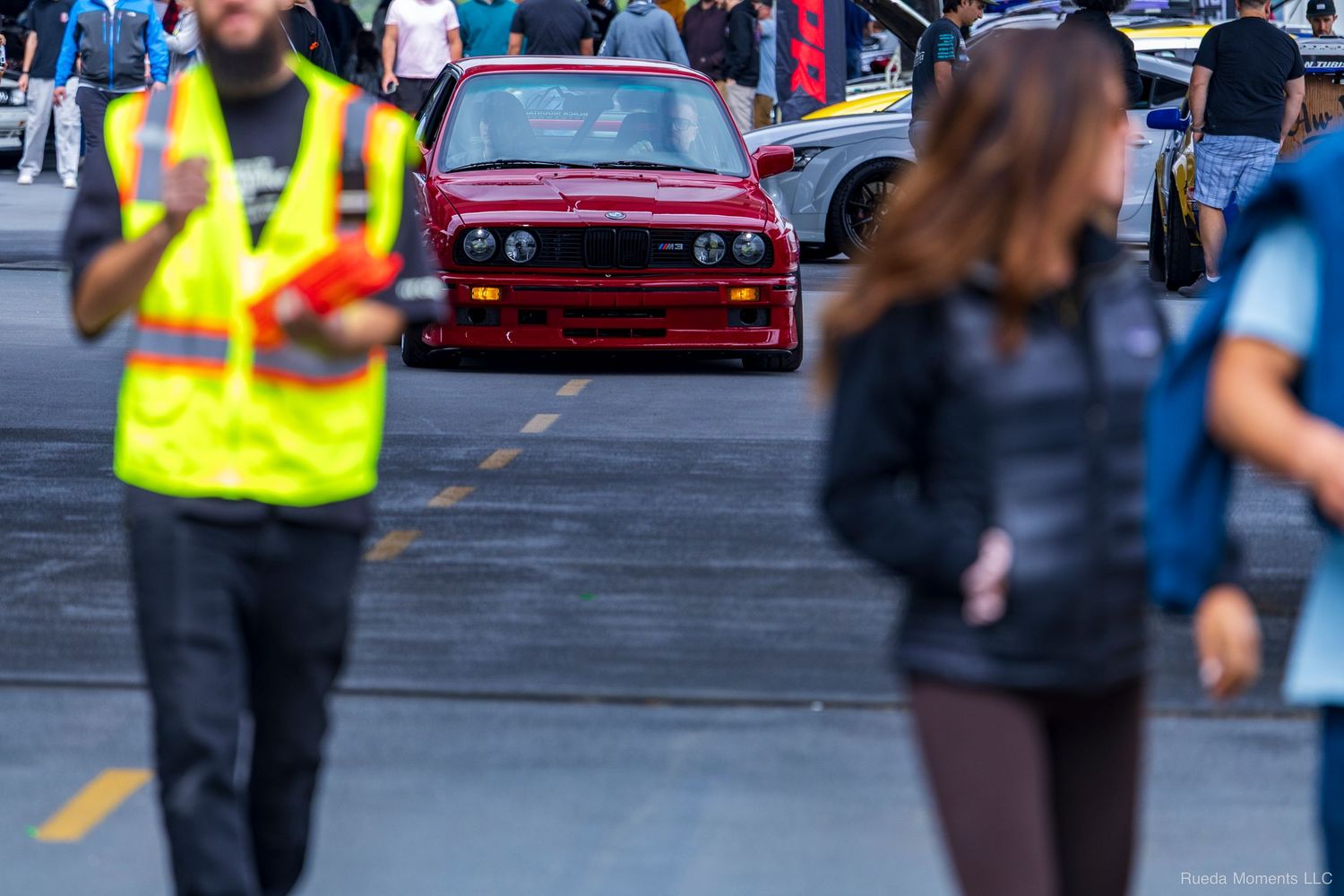 A red BMW E30 M3 drives through a crowded car show event with spectators and staff.