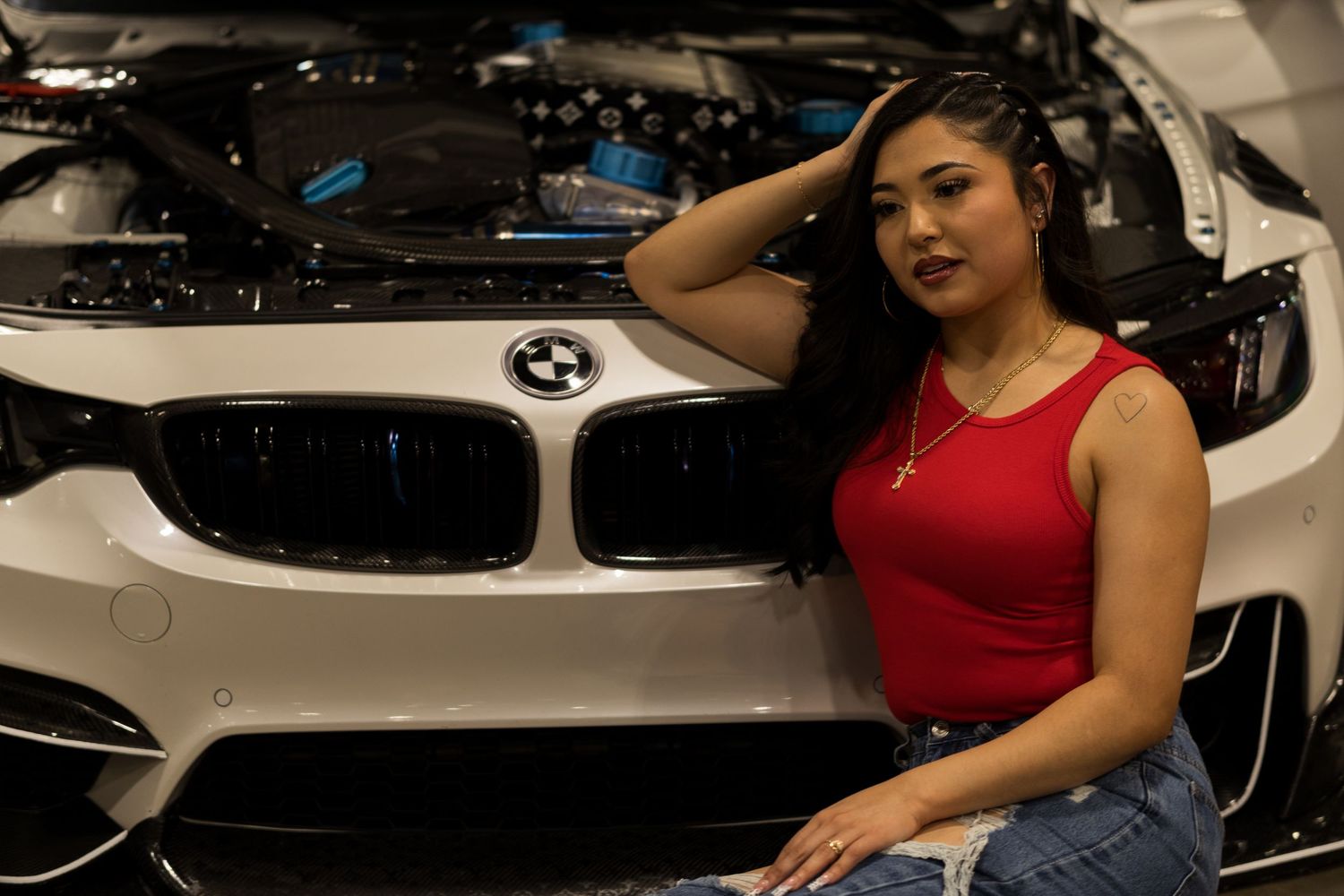 Woman in red top poses beside a white BMW with open hood at a car show.