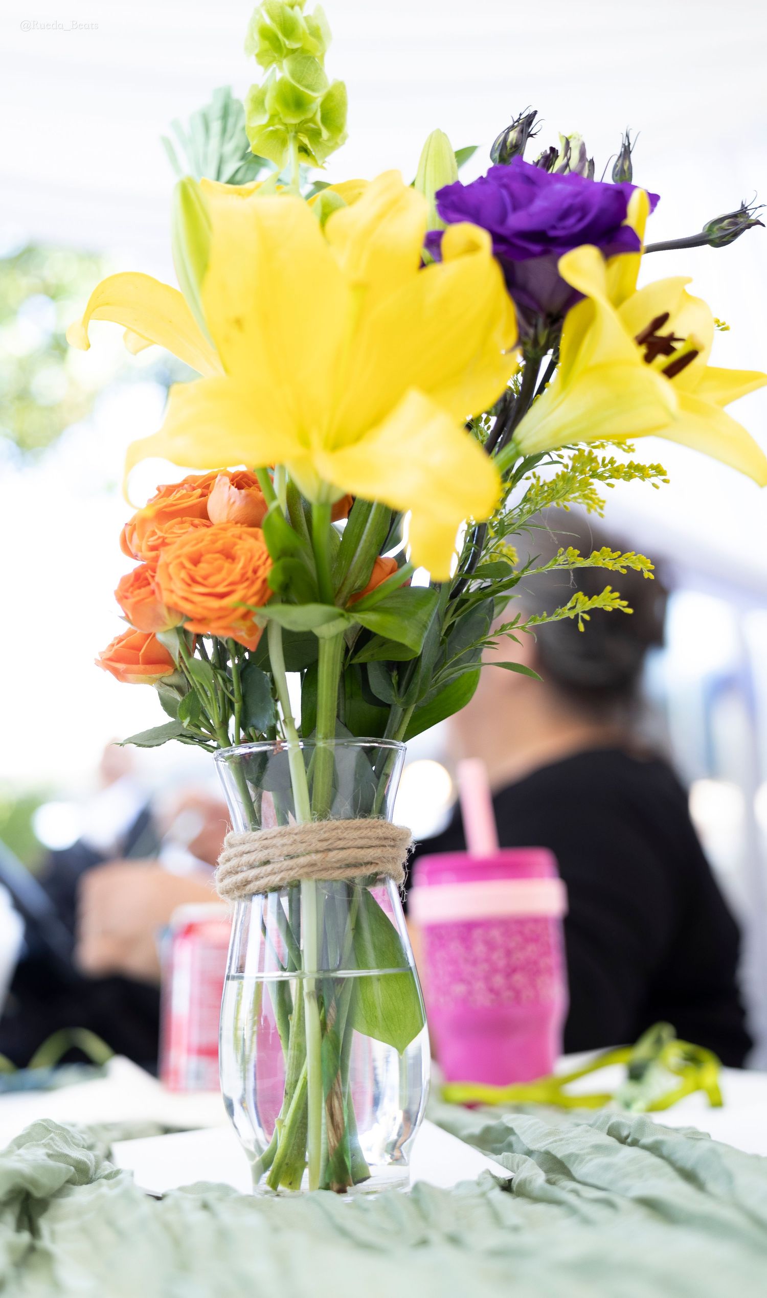 Glass vase with yellow lilies, orange roses, and purple flowers tied with twine on a table at an outdoor event.