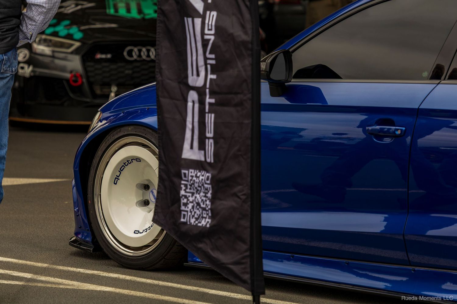 Blue car on white deep dish wheels with a sponsor banner at a car show.