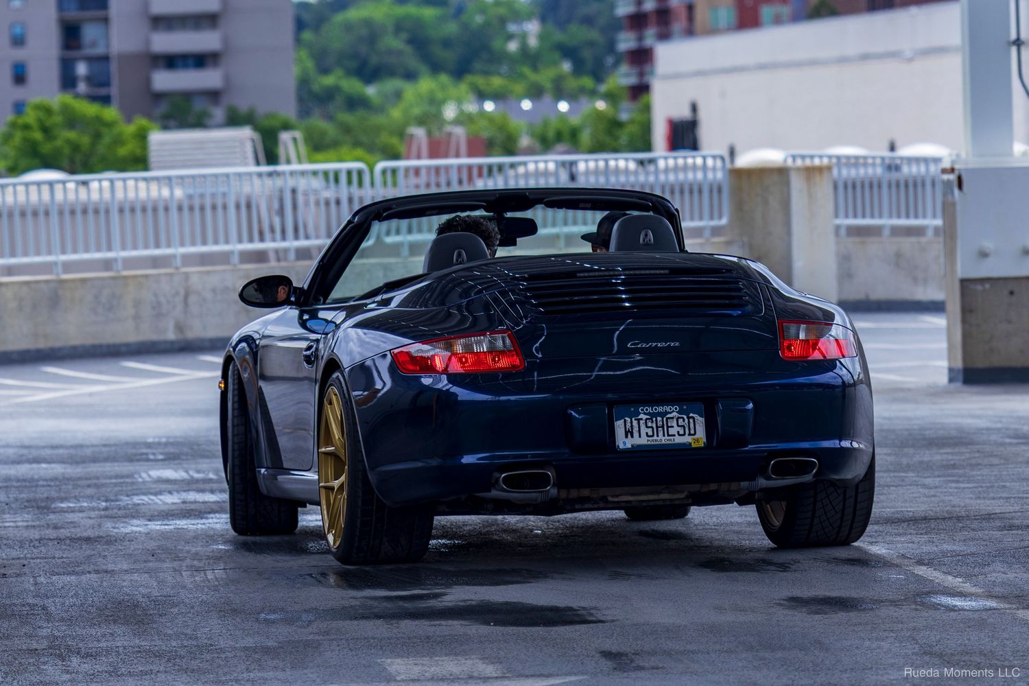 A dark blue Porsche 911 convertible with gold wheels parked on a wet parking structure.