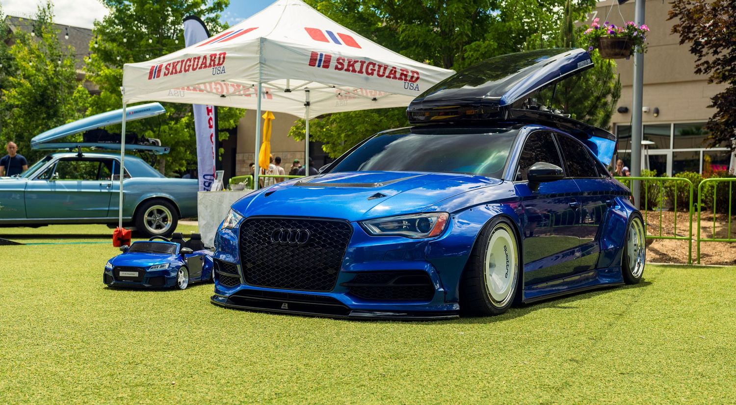 Lowered blue Audi RS3 with roof cargo box on green grass at a car show near a SkiGuard tent.