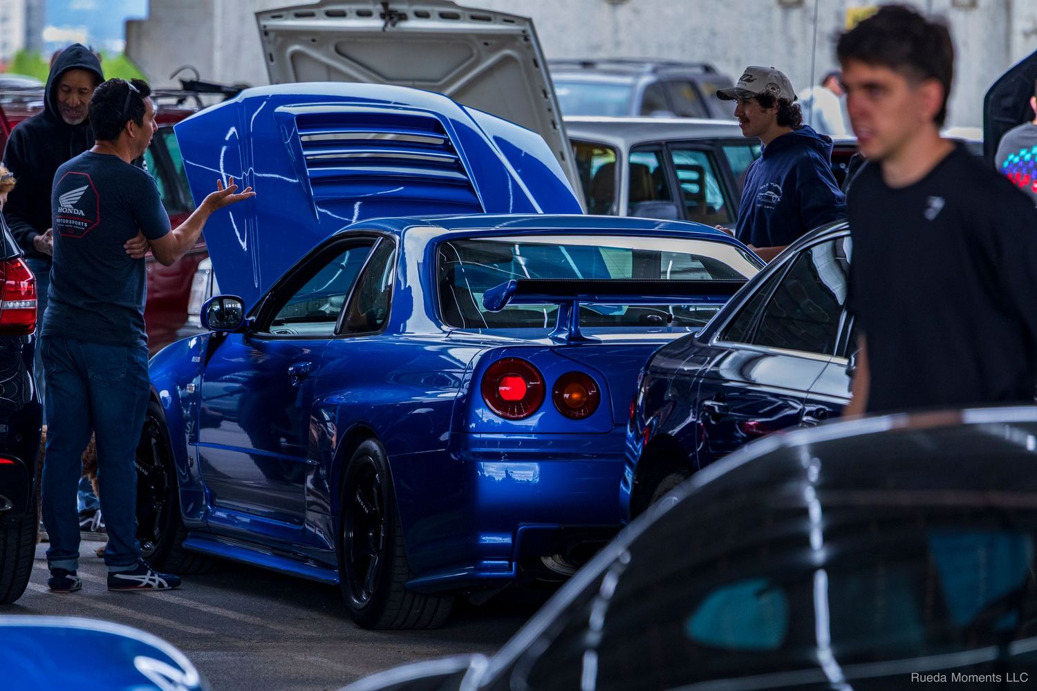 Blue Nissan Skyline GT-R R34 with hood open at a car meet surrounded by enthusiasts.