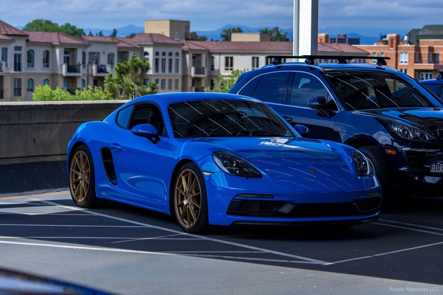 Striking blue Porsche 718 Cayman with gold wheels parked at an outdoor car show.