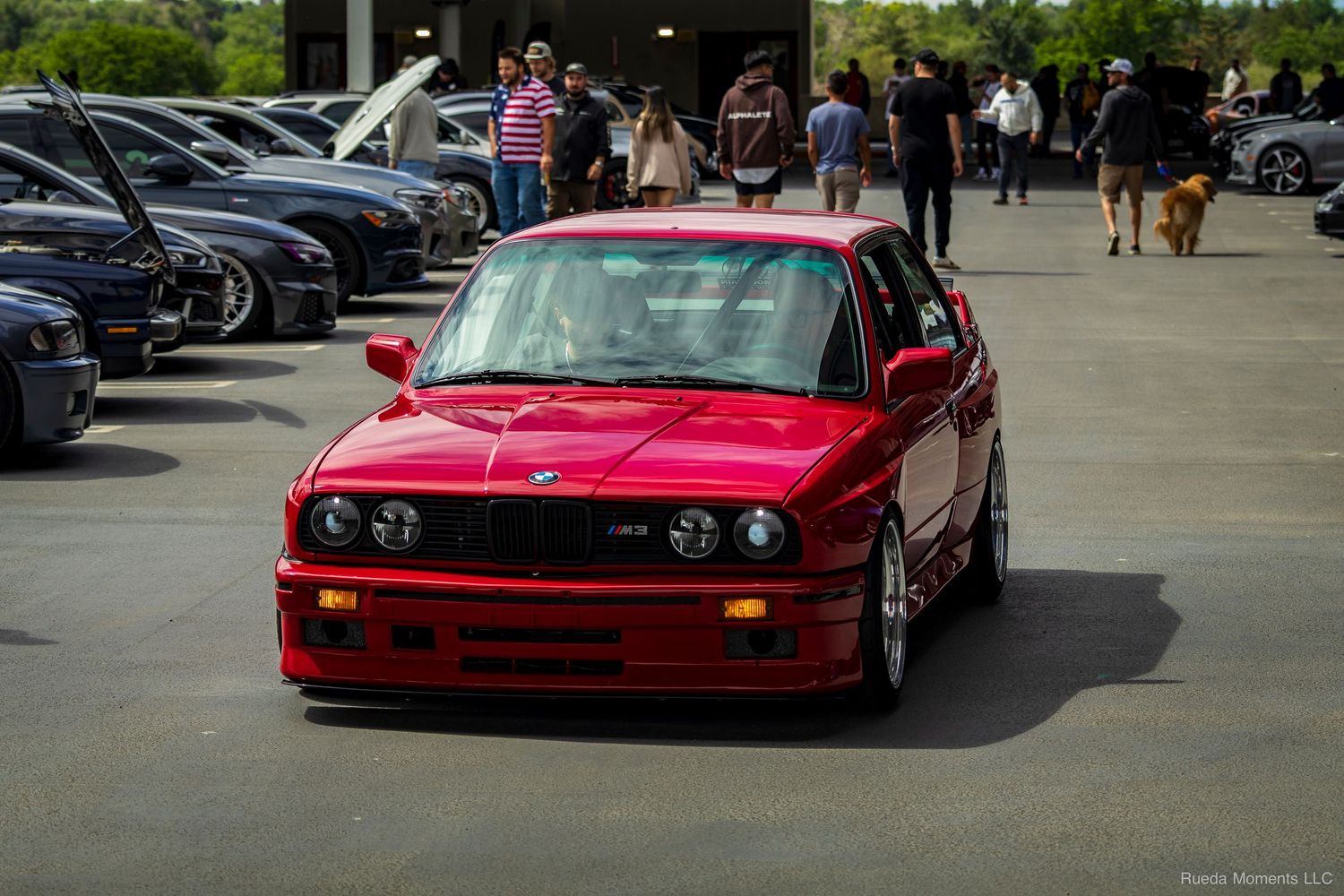 Red BMW E30 M3 at a car show with spectators and other vehicles in the background.