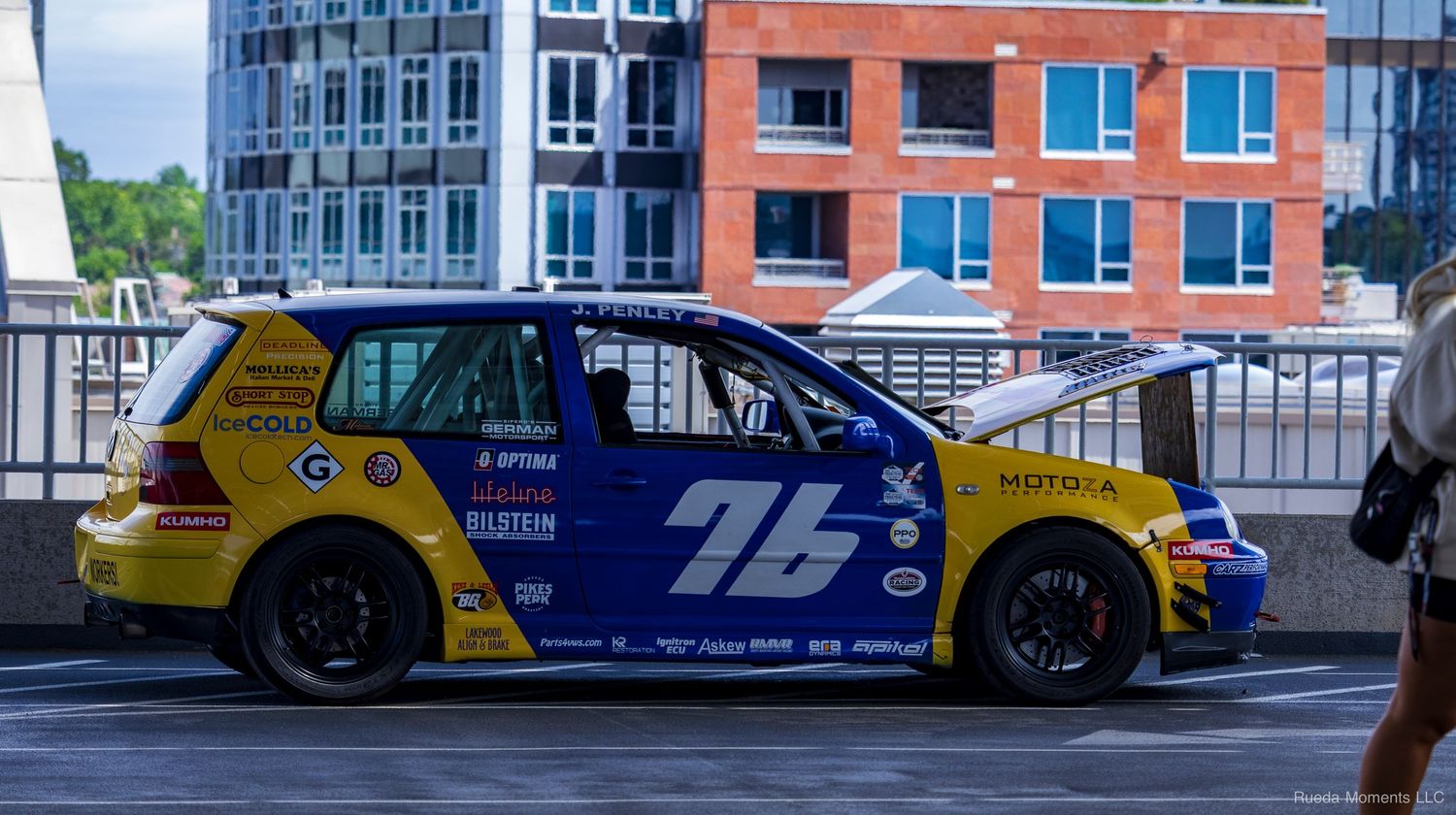 Blue and yellow VW Golf race car number 76 with sponsor decals parked on a street near urban buildings.