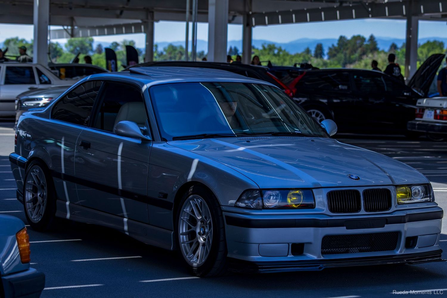 A silver BMW E36 M3 with wide body kit and mesh wheels at an outdoor car show.