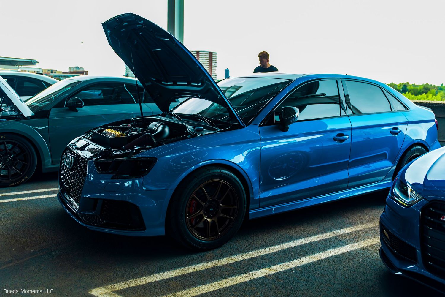 Blue Audi S3 sedan with hood open on display at a sunny outdoor car show event.