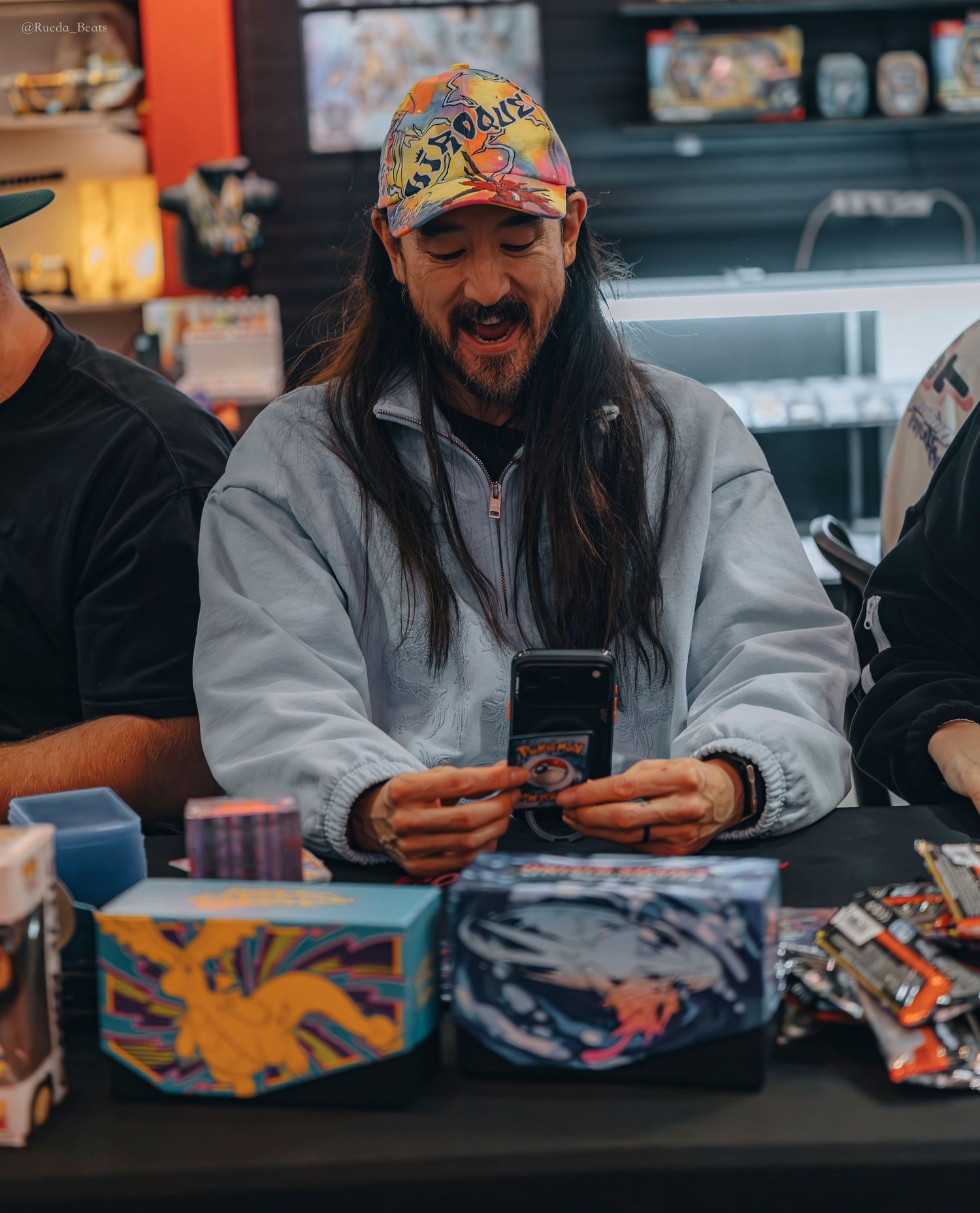 Smiling long-haired man in tie-dye cap excitedly opening Pokemon trading card packs at a store event.
