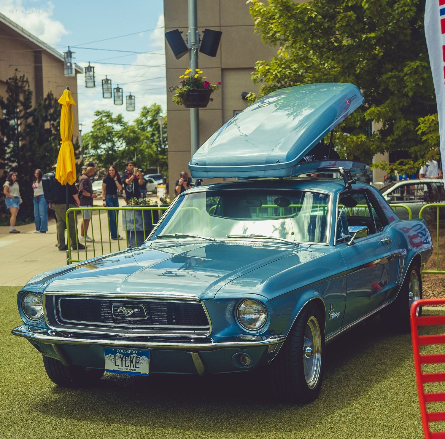 Classic blue Ford Mustang with roof cargo box displayed at an outdoor car show on a sunny day.