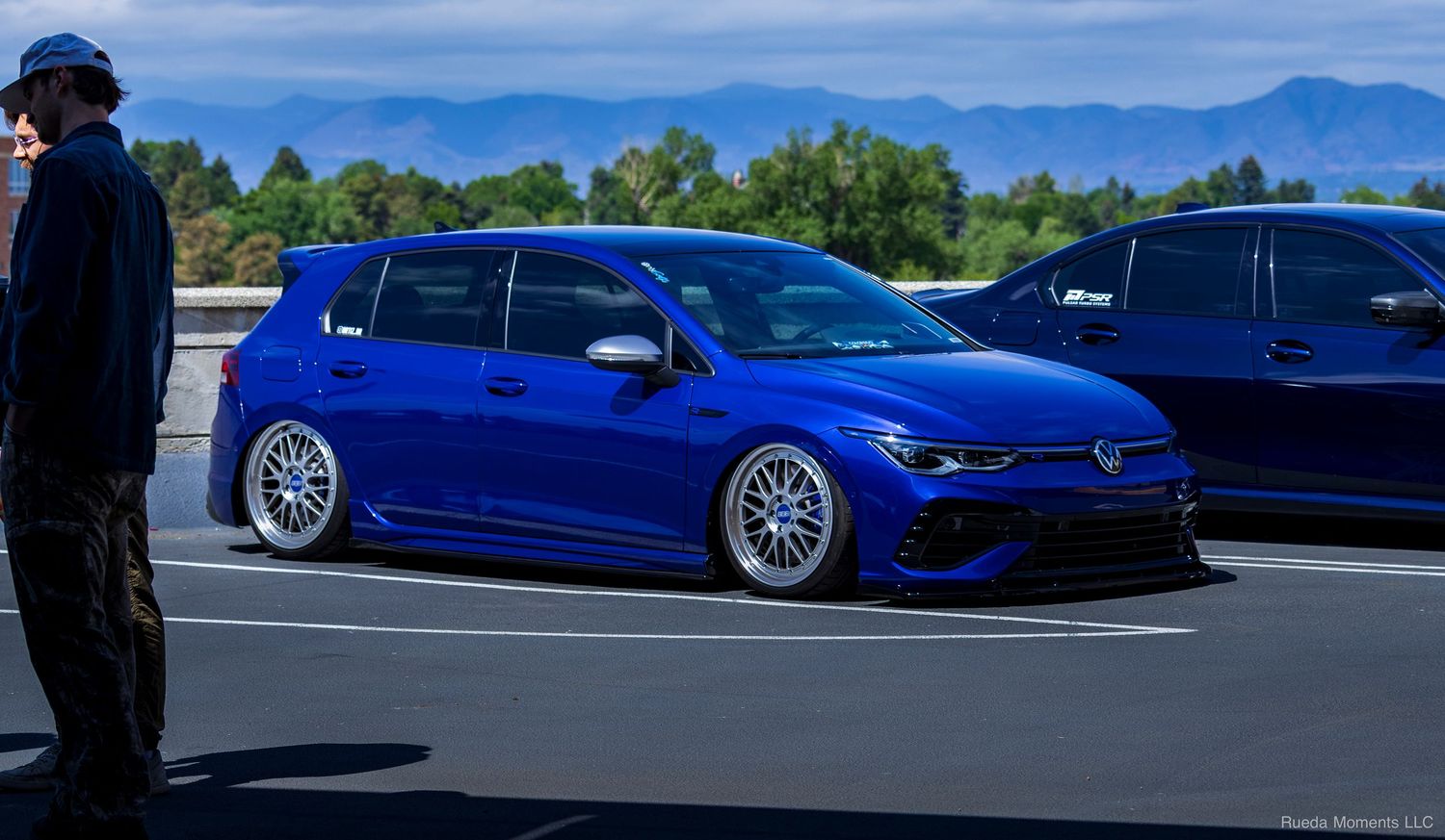 Slammed blue Volkswagen Golf R on BBS wheels parked at an outdoor car meet with mountains.
