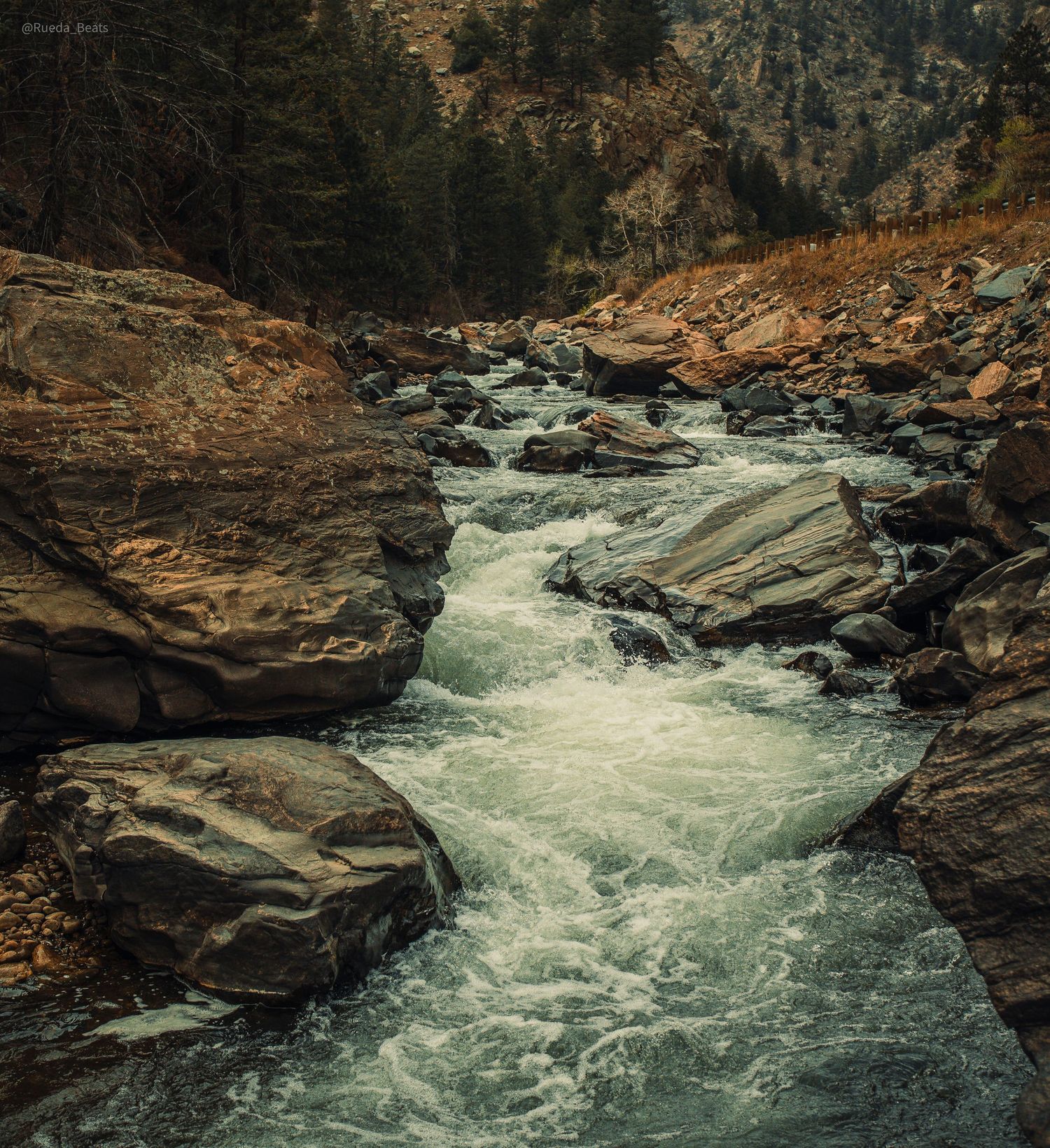 Scenic mountain river with rushing white water rapids winding through large boulders surrounded by pine forest and rocky terrain.