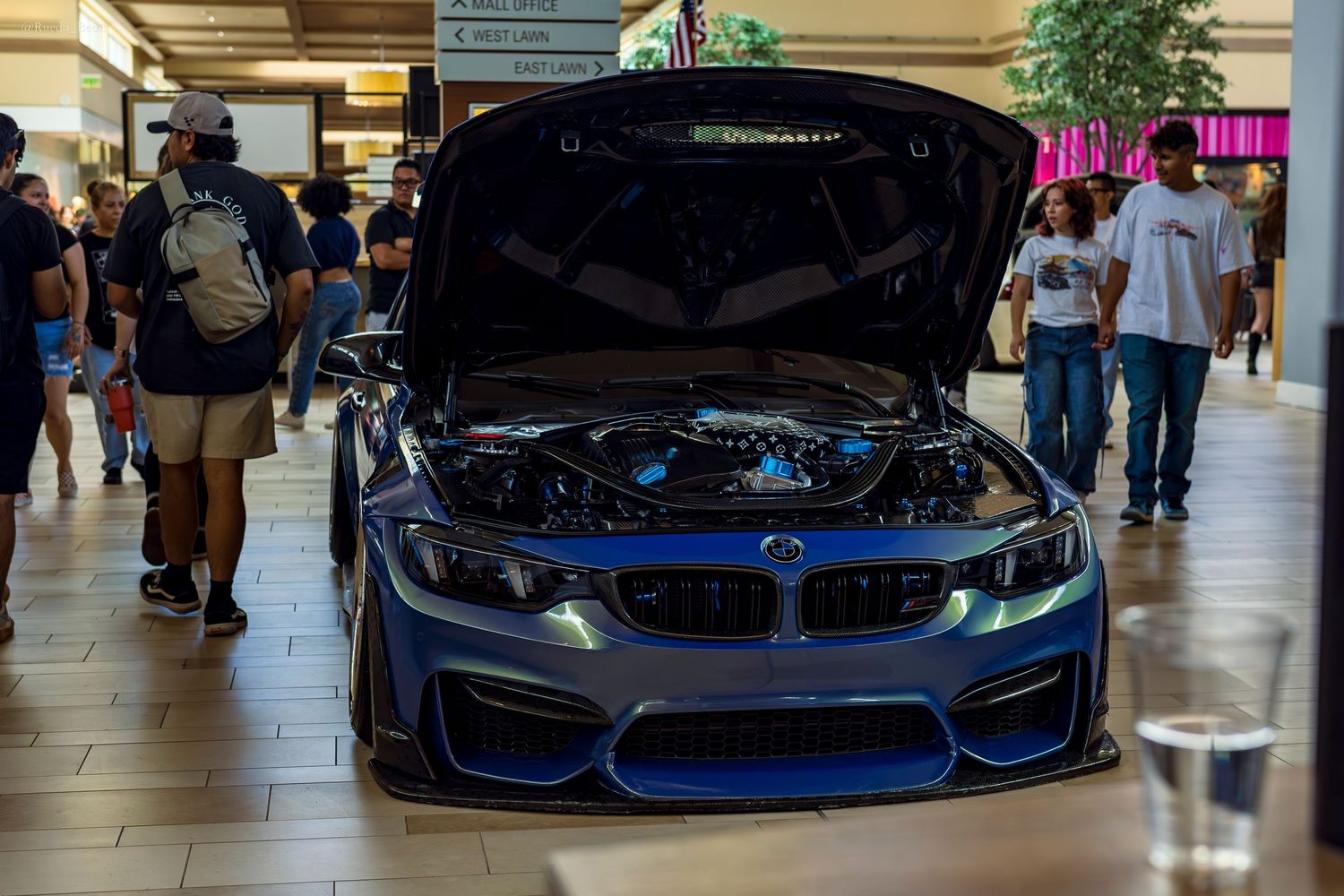 A customized blue BMW M3 with open hood showcased at an indoor shopping mall car show.