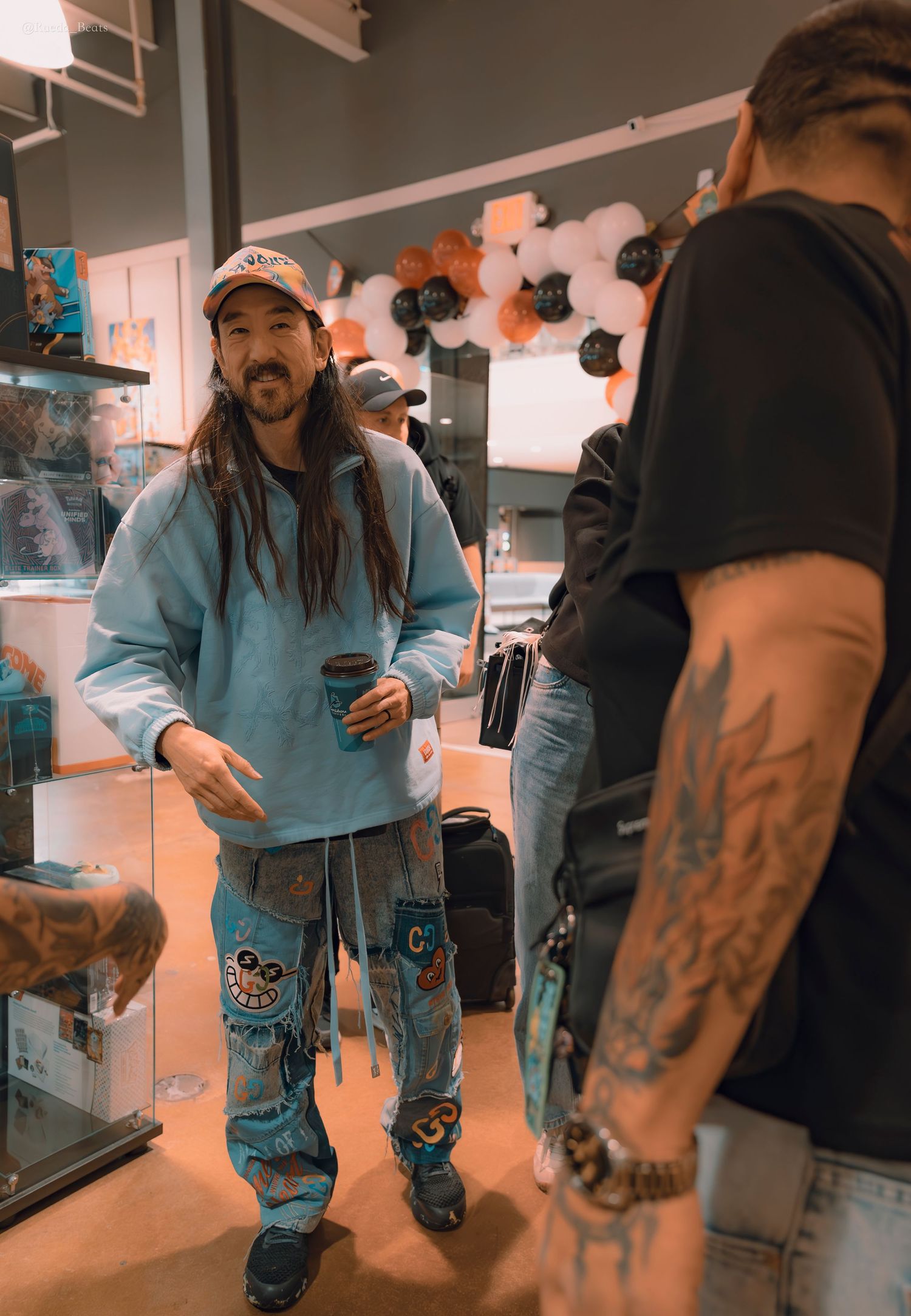 Man with long dreadlocks wearing light blue hoodie and patched jeans holding coffee in a retail store.