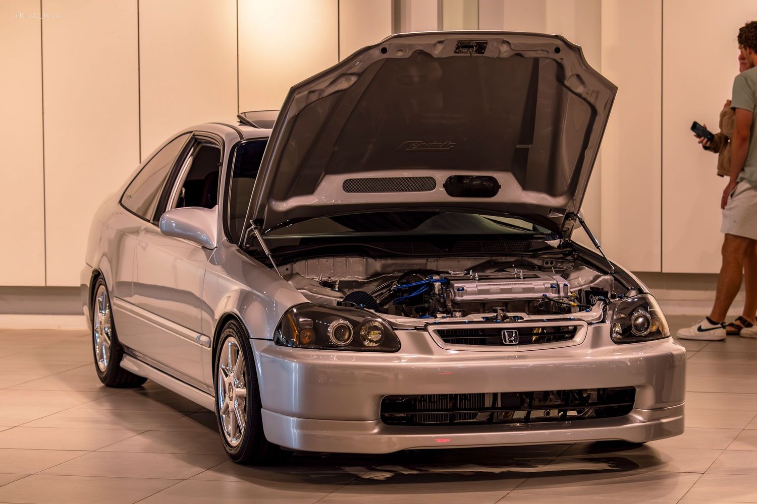 Silver Honda Civic EK with hood open revealing engine bay at an indoor car show.
