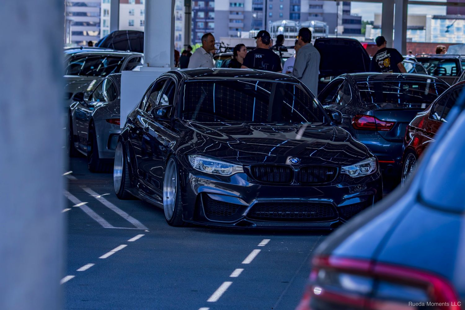 Modified black BMW M5 with wide body kit parked in a covered parking garage car show display.