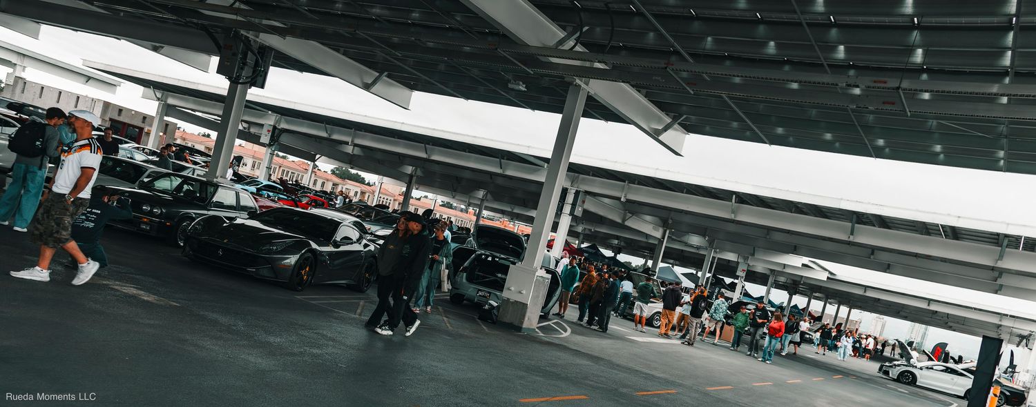 Wide panoramic shot of exotic and modified cars gathered under a covered parking structure.