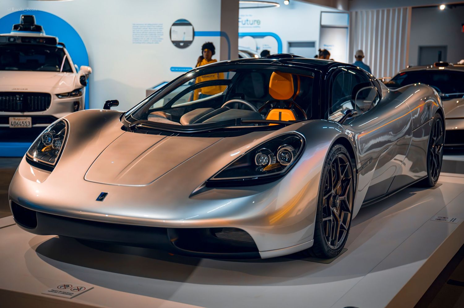 Silver Gordon Murray T.50 supercar on display at an auto show with orange interior visible.