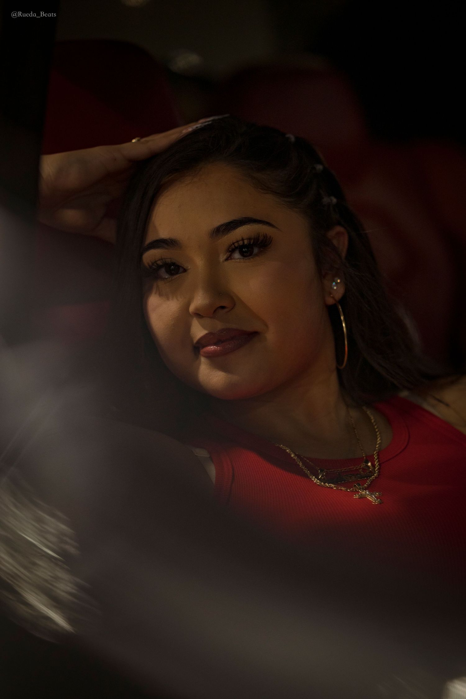 Young woman in red top with layered gold necklaces and hoop earrings poses in dramatic low-key lighting.
