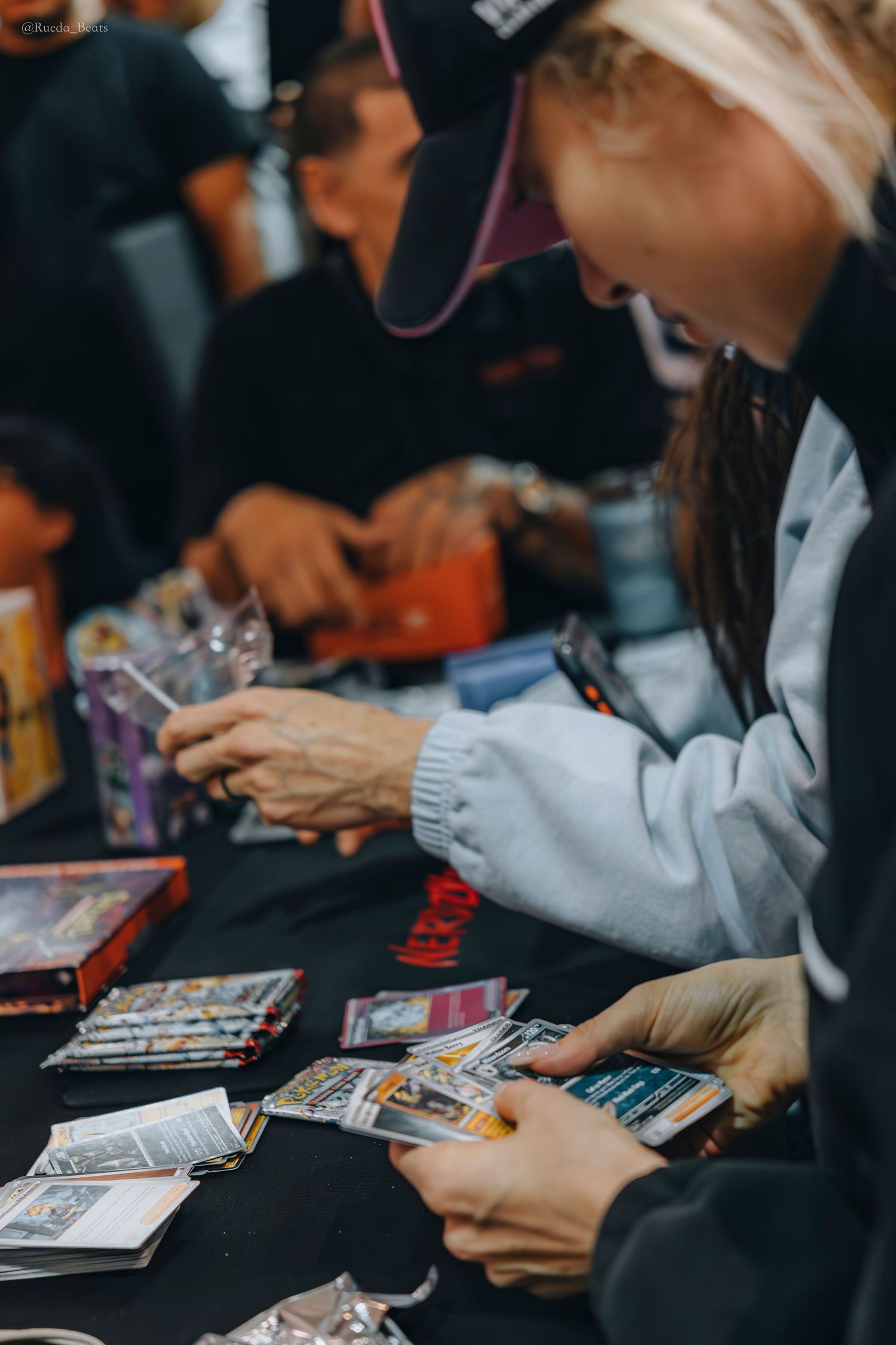 Blonde woman sorting through Pokemon trading cards at a black table during a card opening event.