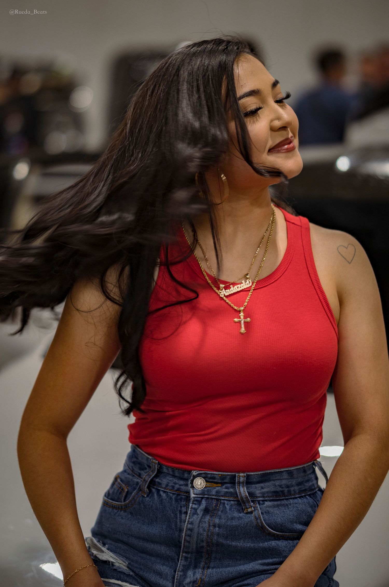 Young woman with flowing dark hair wearing a red tank top and denim shorts with a gold cross necklace.