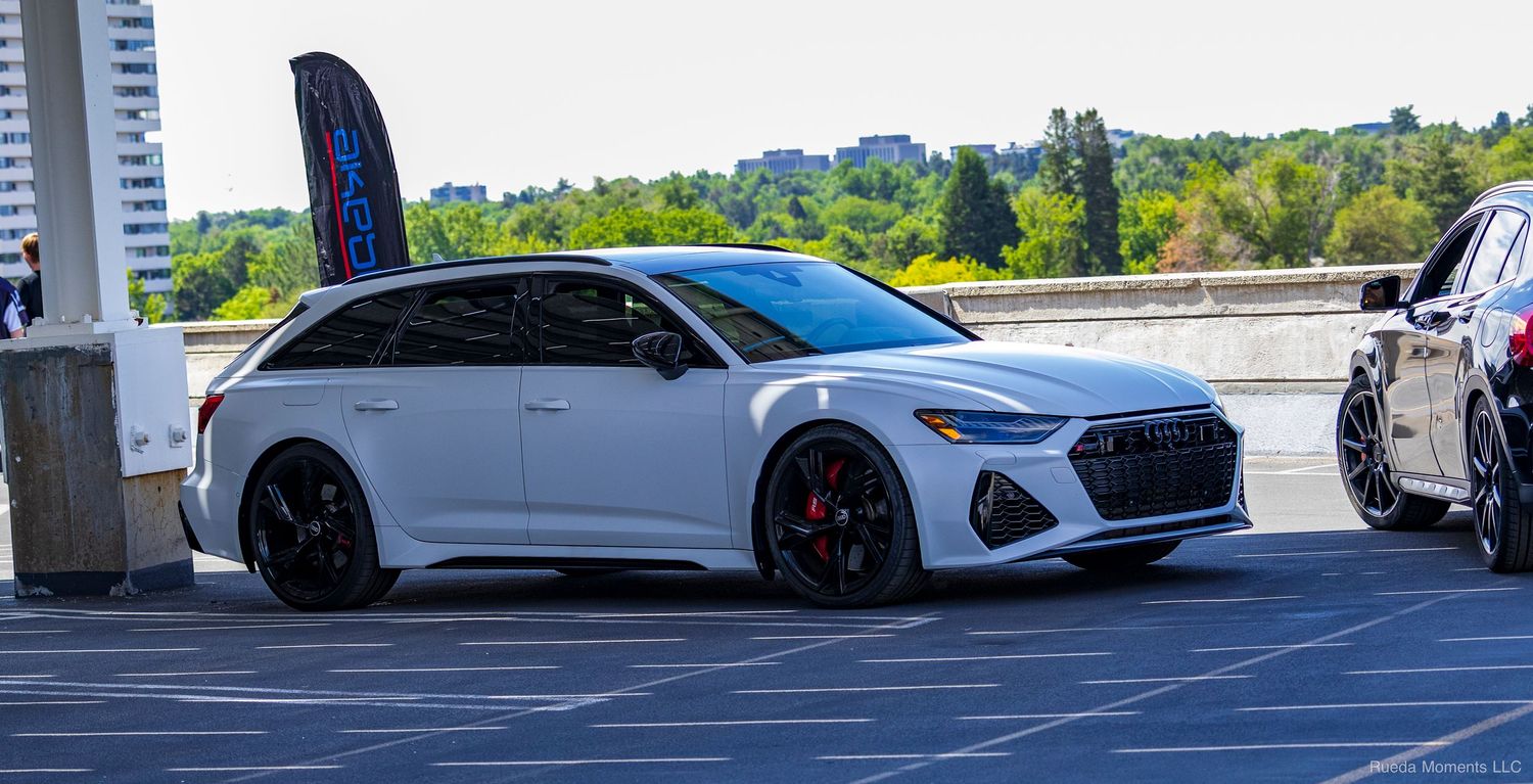 White Audi RS6 Avant wagon with black wheels parked in a lot at an outdoor car show.