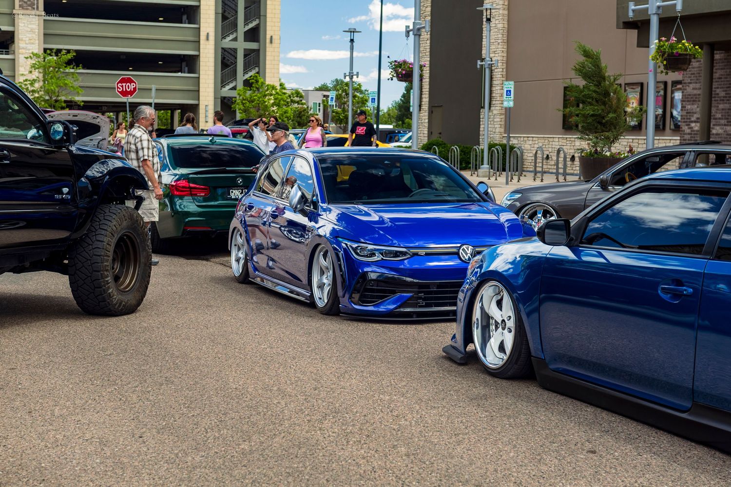 Two blue lowered VW Golf cars parked at an outdoor car show on a sunny day.