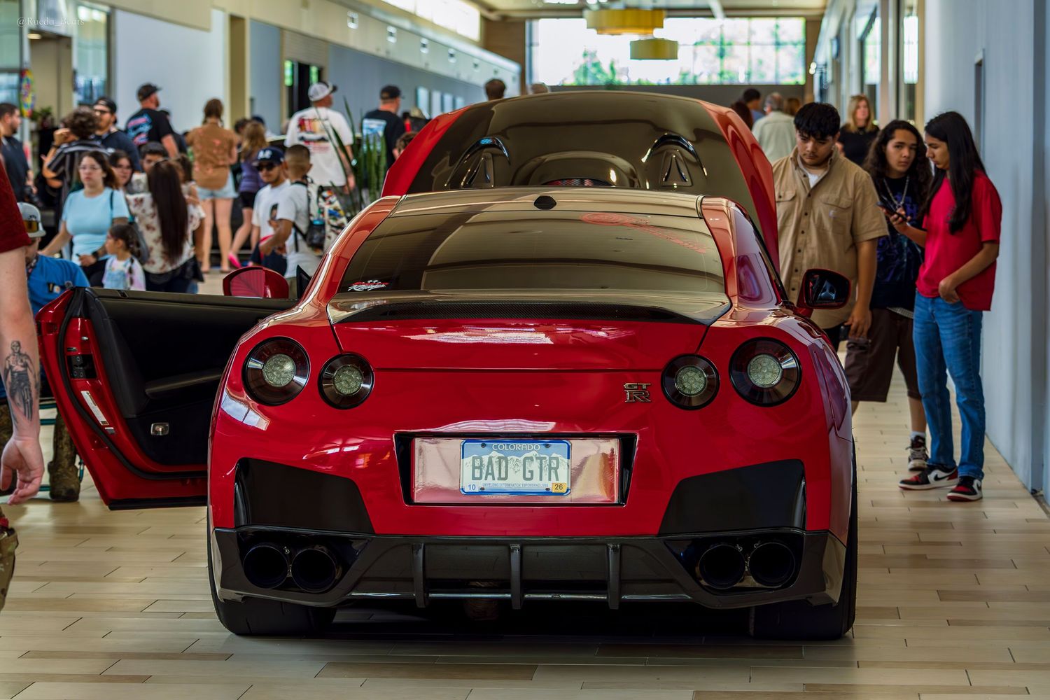 Modified red Nissan GT-R with scissor doors open viewed from rear at crowded car show.