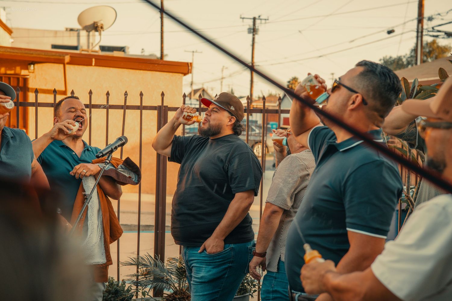 Group of men laughing and drinking outdoors at a casual social gathering near a fence during golden hour.