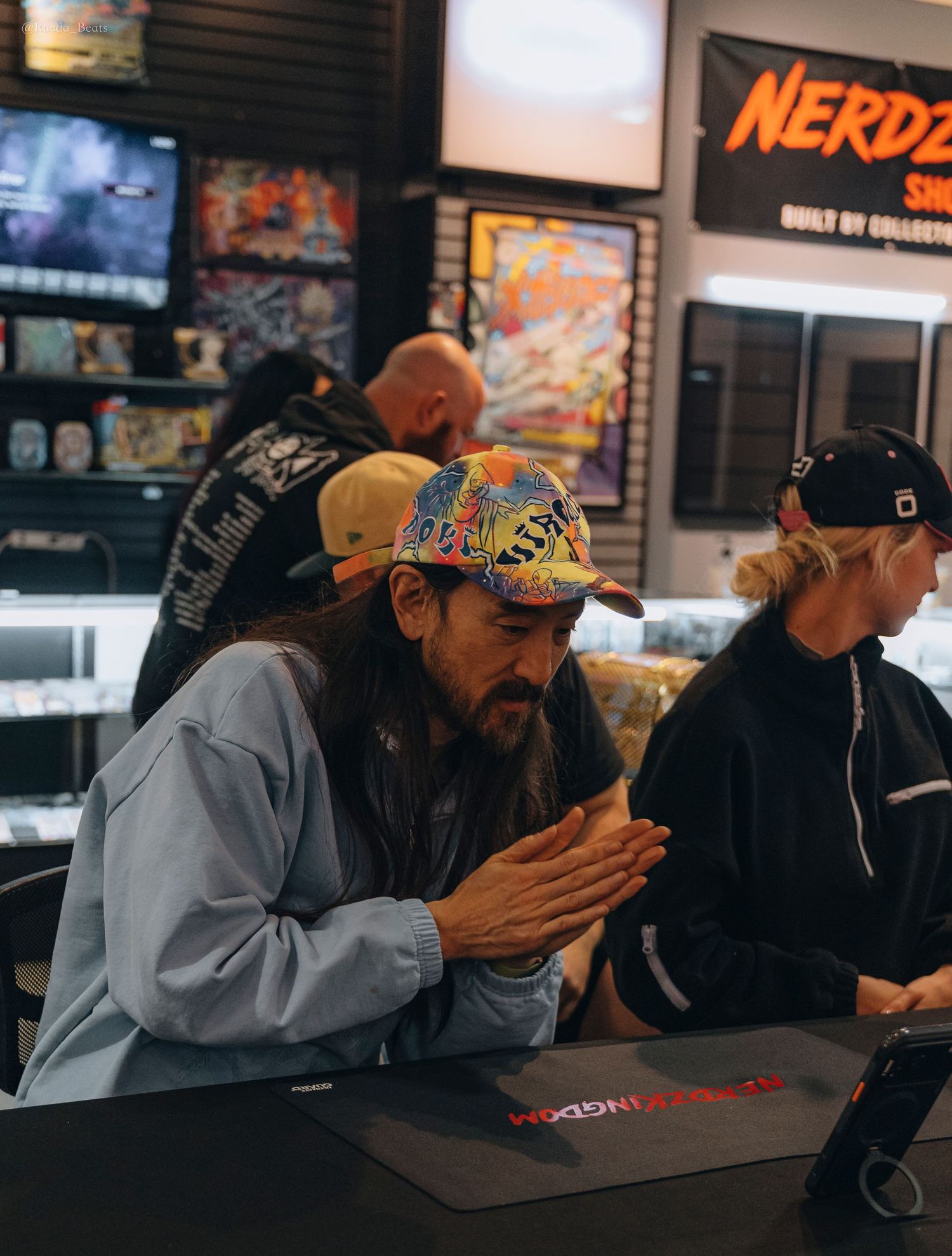 Man in colorful cap with hands clasped at Nerdz Kingdom collectibles store counter.