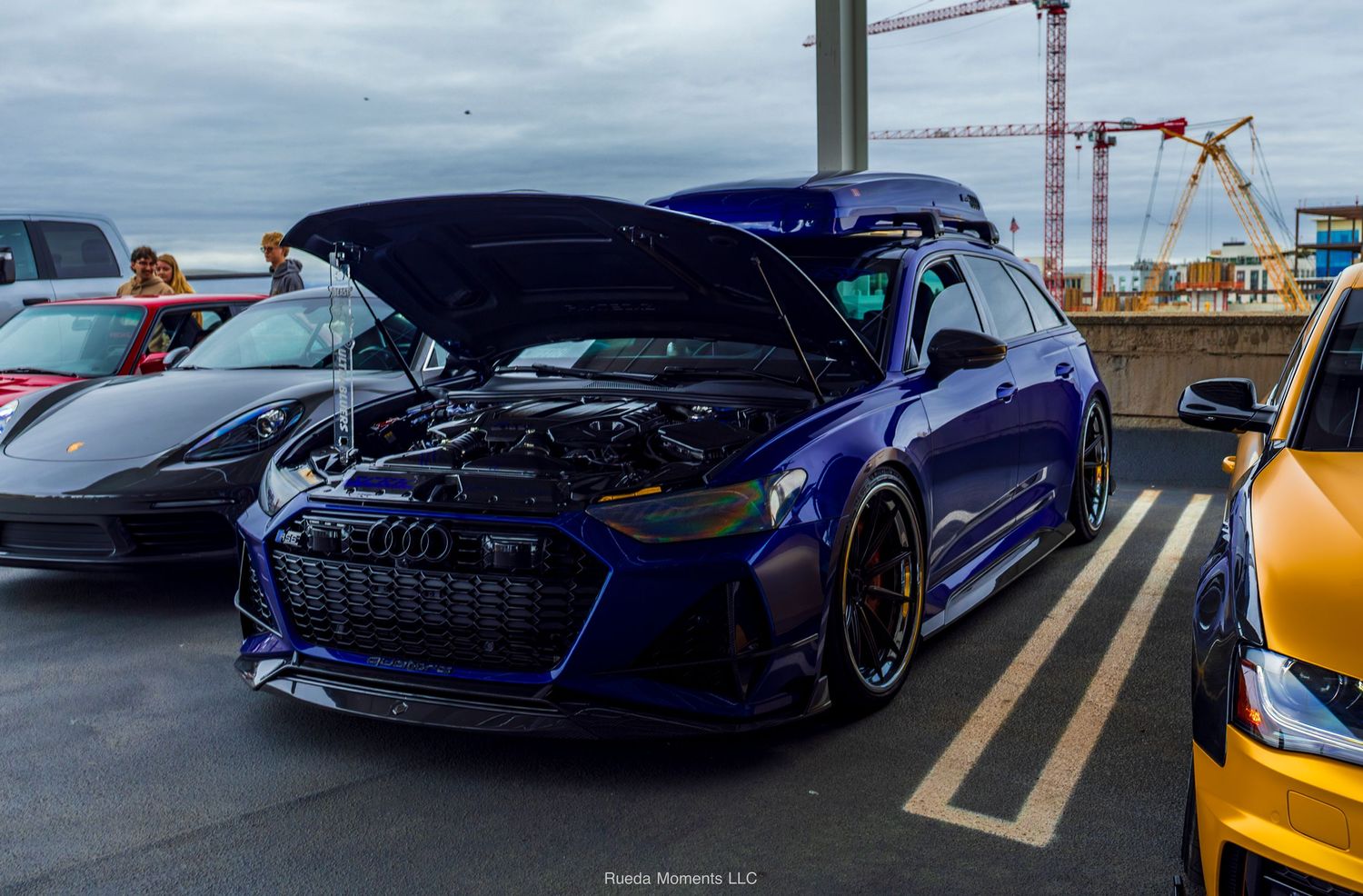 Widebody blue Audi RS6 with open hood at outdoor car show.