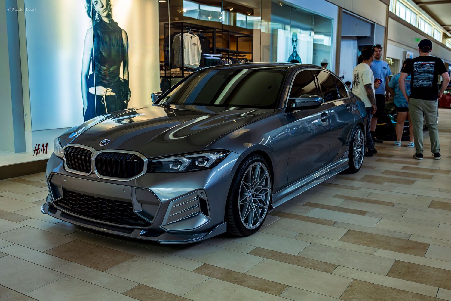 A modified gray BMW 3 Series with wide body kit and custom wheels at an indoor car show.