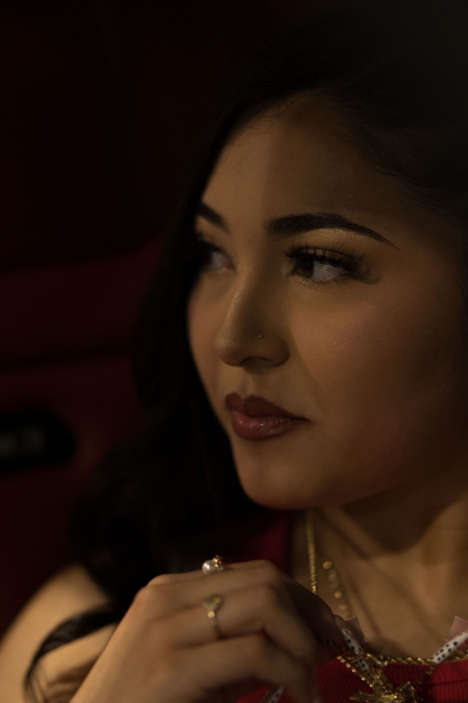 Close-up portrait of a young woman with dark hair, wearing a ring and gold necklace, gazing thoughtfully.