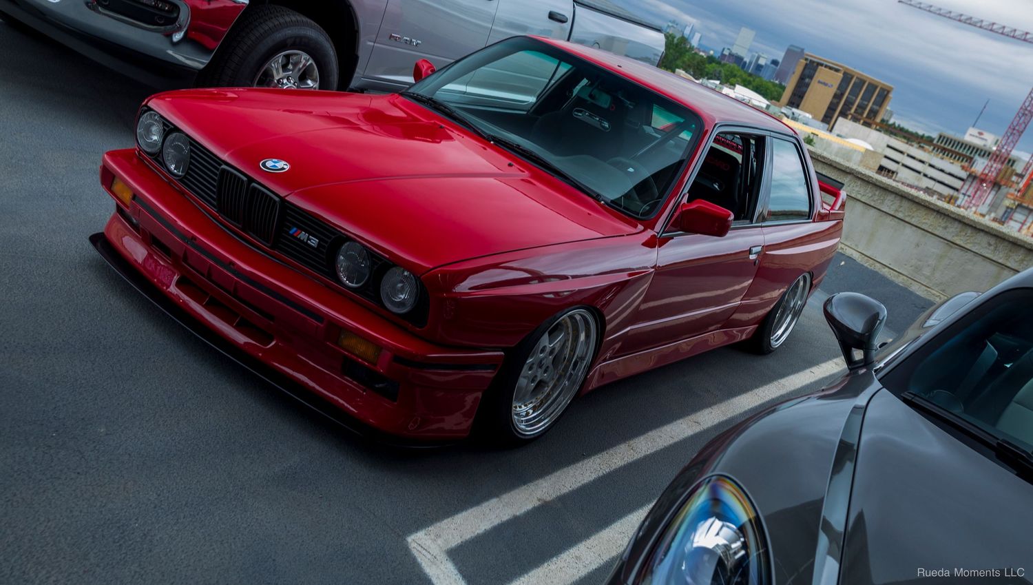 Classic red BMW E30 M3 on BBS wheels parked at an outdoor automotive car meet event.
