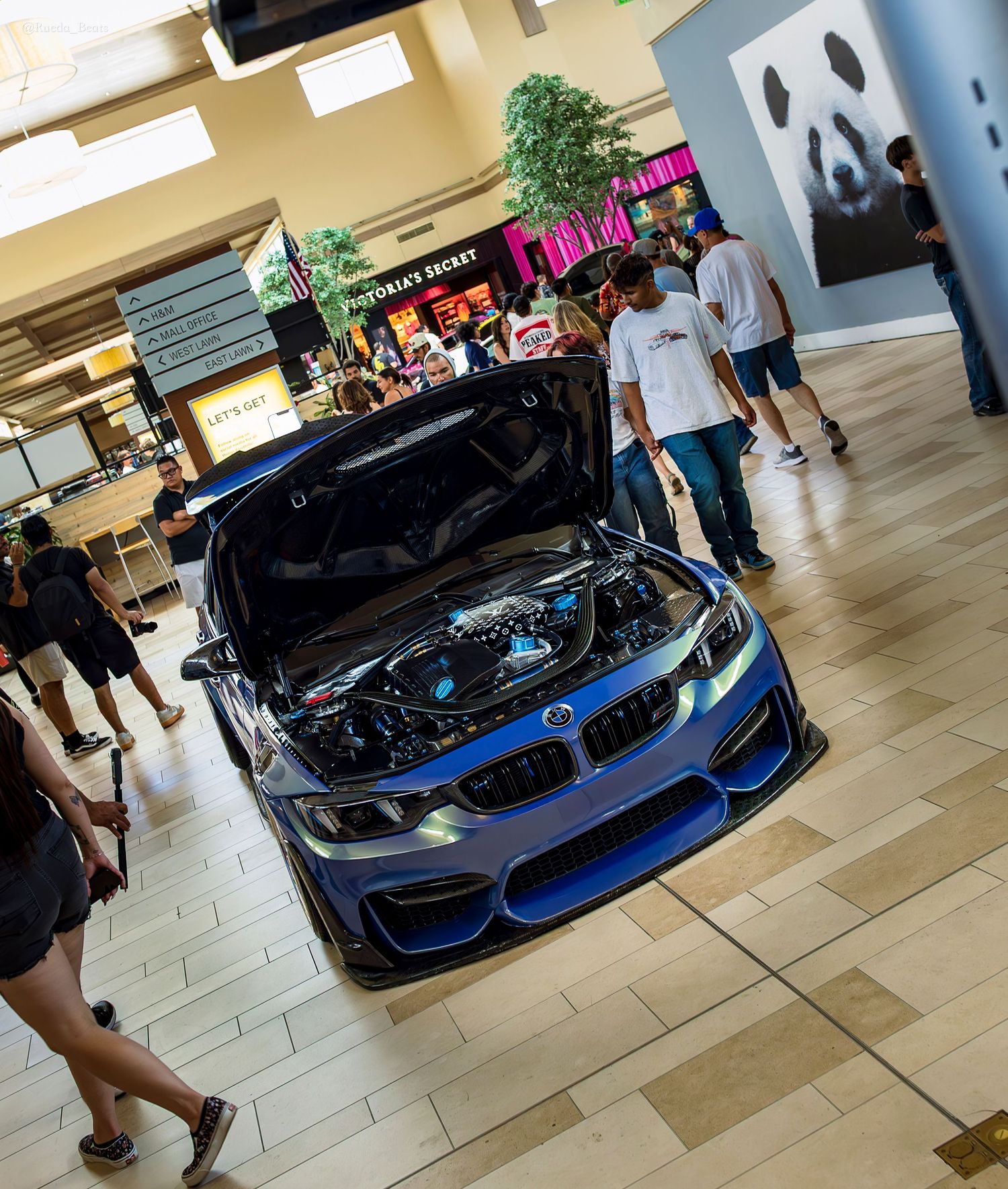 A modified blue BMW with hood open on display inside a shopping mall with spectators walking around.