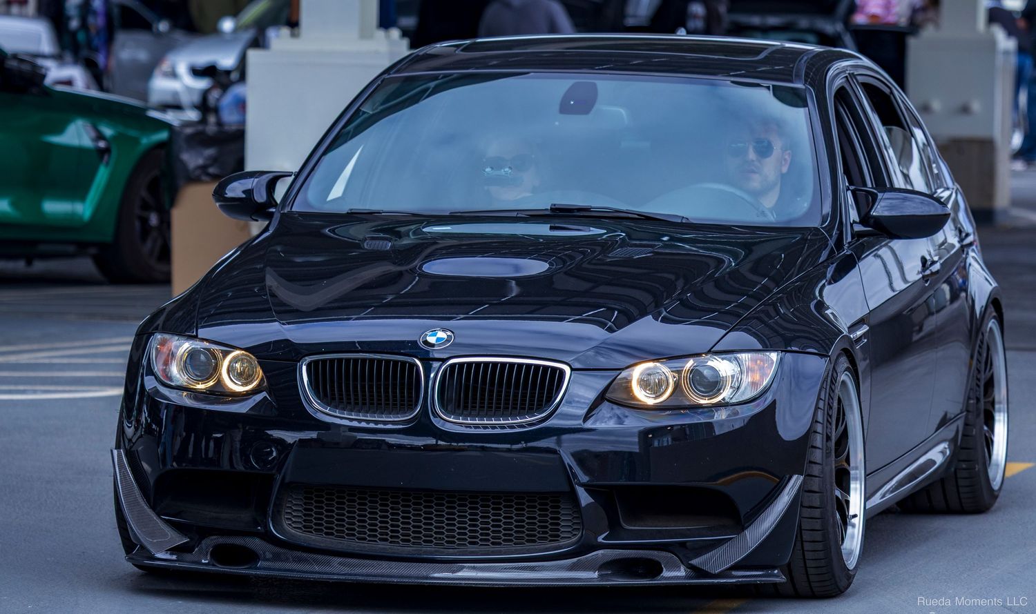 A black modified BMW M3 E90 with angel eyes and wide body kit at a car show.