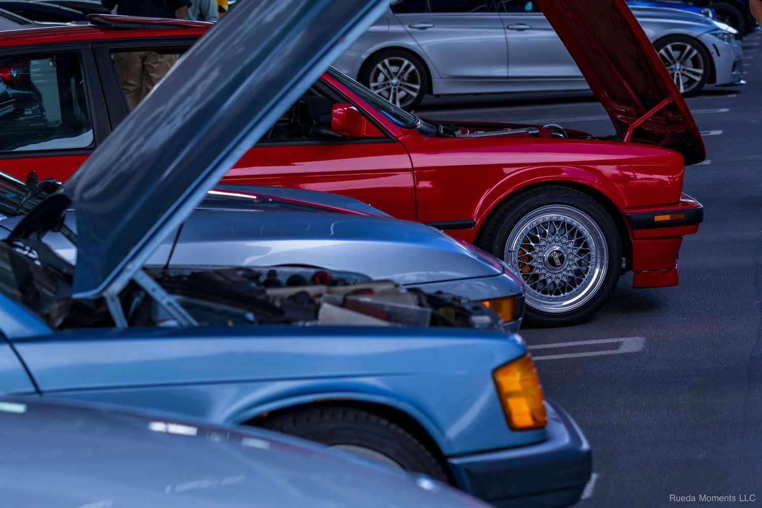 Classic BMW E30 and vintage cars with hoods open at a twilight car show event.