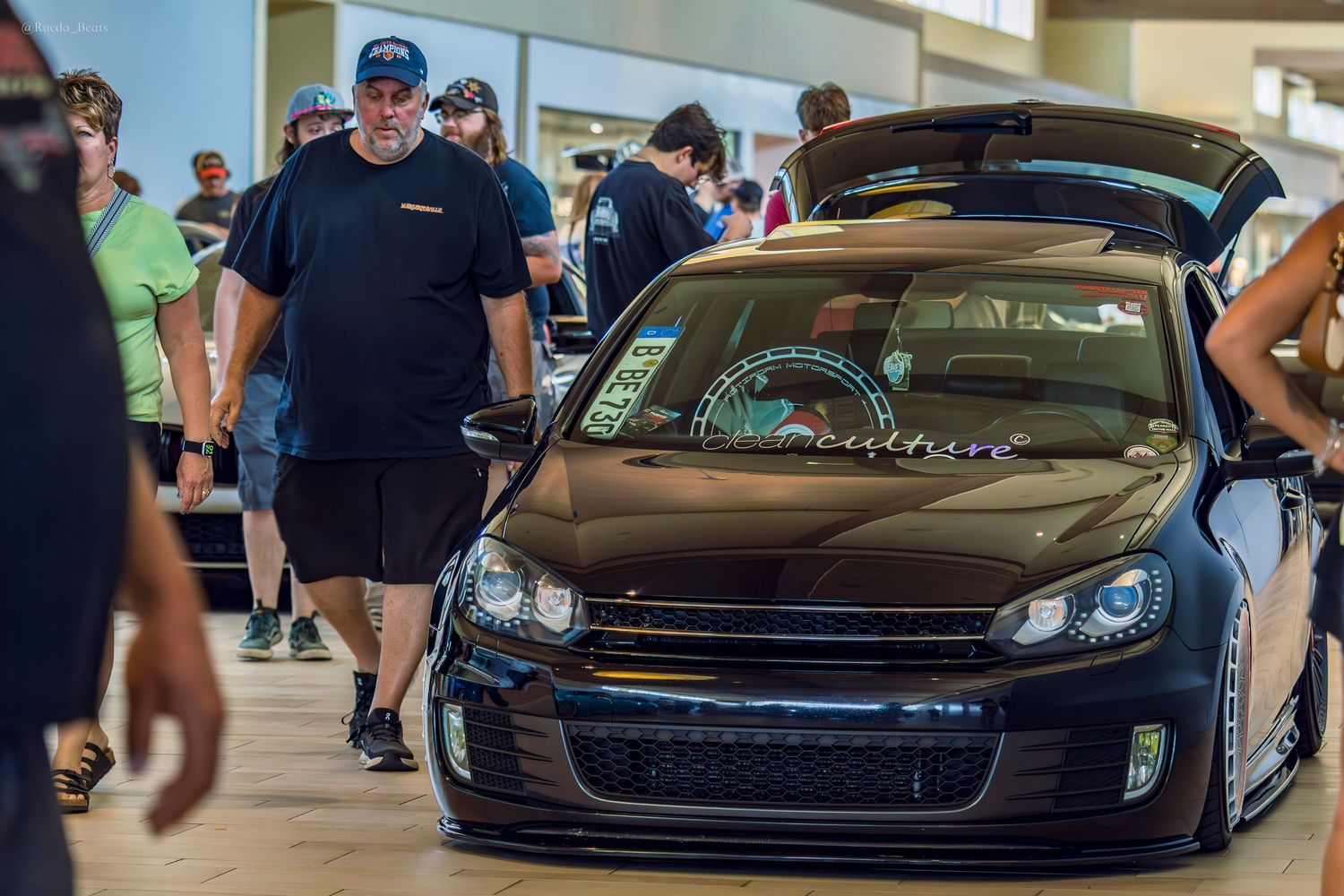 Slammed black Volkswagen Golf MK6 with gull-wing doors open at indoor car show event.