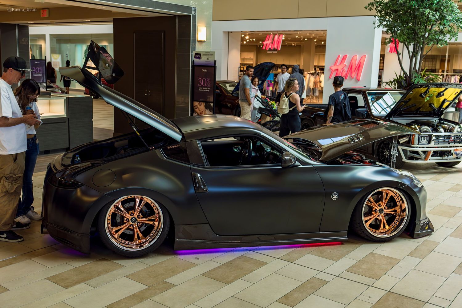 A matte black Nissan 370Z with copper wheels and purple underglow on display inside a mall.