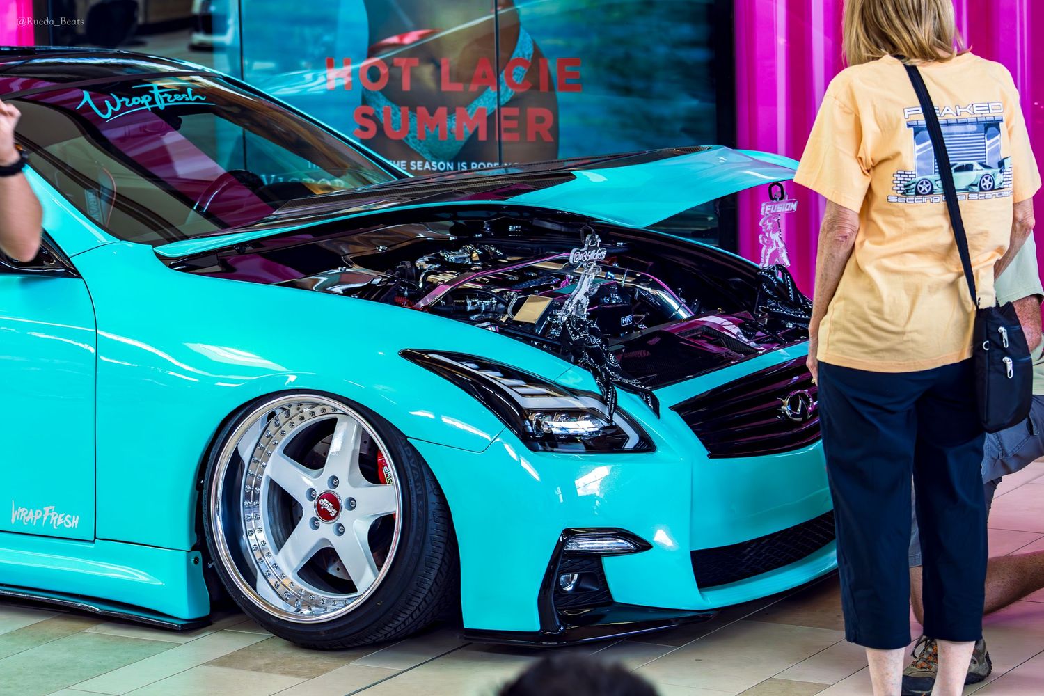 Teal bagged Infiniti G37 with hood open and custom wheels at an indoor car show event.
