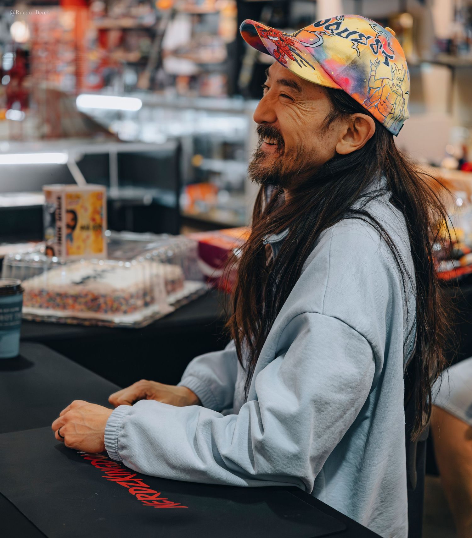 Long-haired man in colorful cap laughing behind counter in retail store.