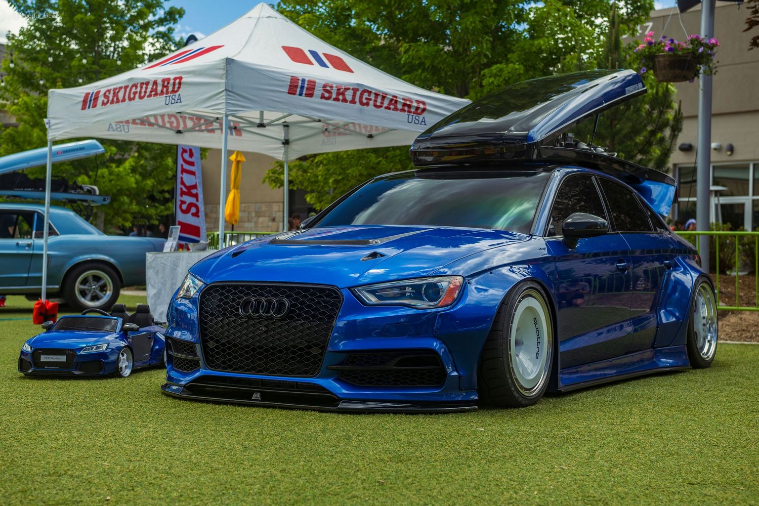 Slammed blue Audi A3 with body kit and roof box parked on grass at a SkiGuard sponsored car show.