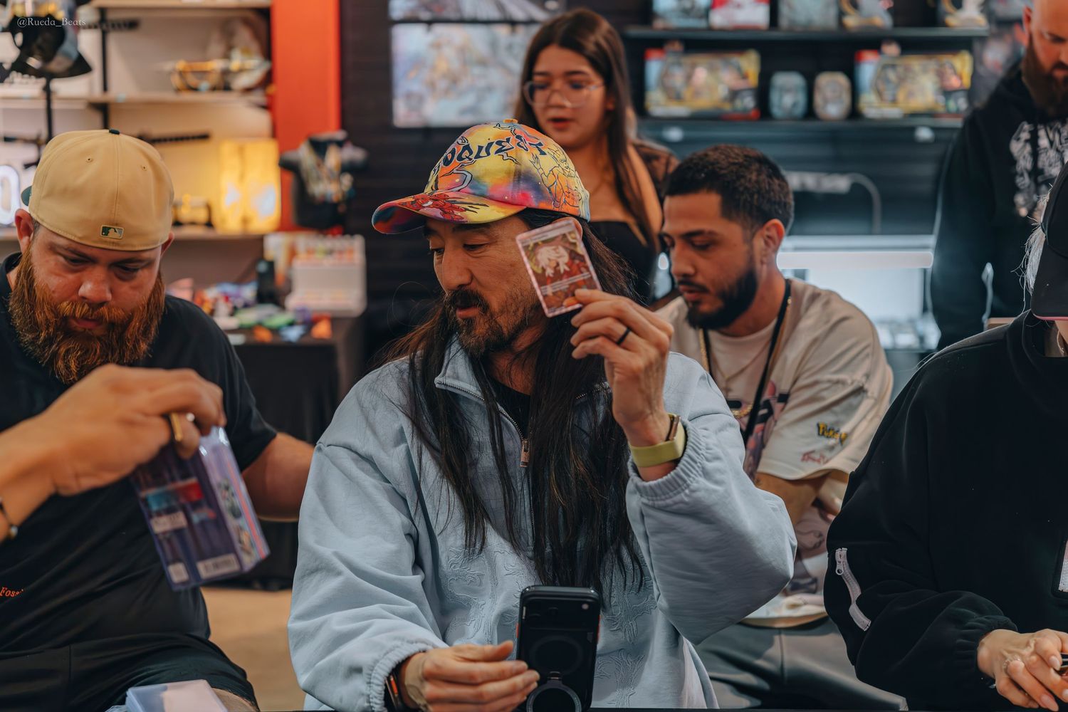 Long-haired man in colorful cap holding up a trading card while seated at a retail store event.