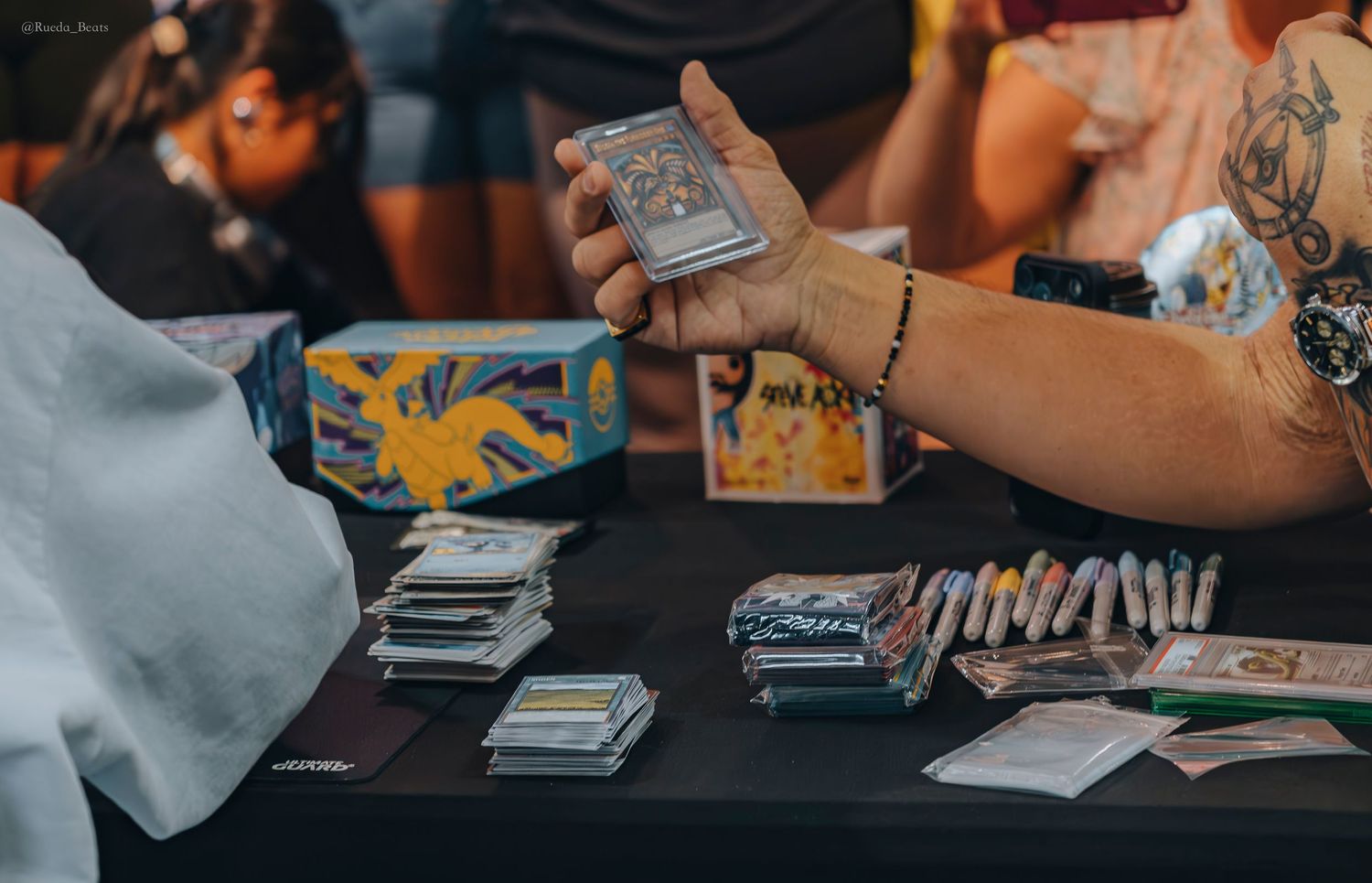Tattooed hand holds trading card deck at collector event table.
