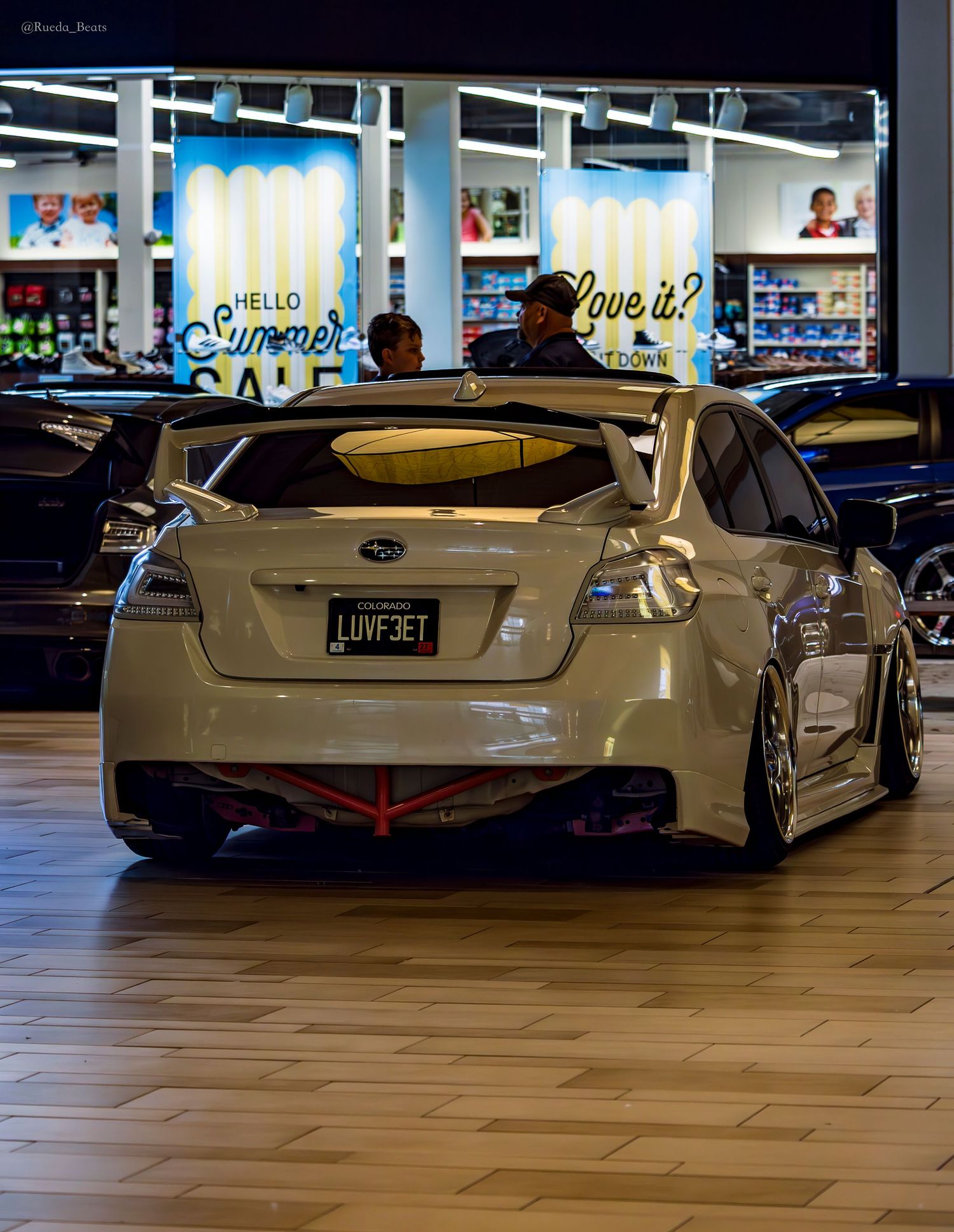 White Subaru WRX STI with large rear wing and LUVF3ET plate at mall car show.