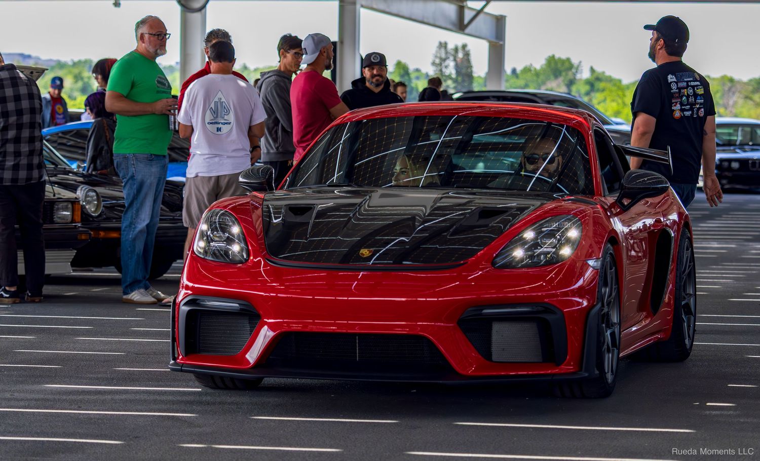 Red Porsche 718 Cayman GT4 at a car show with enthusiasts gathered in a parking lot.
