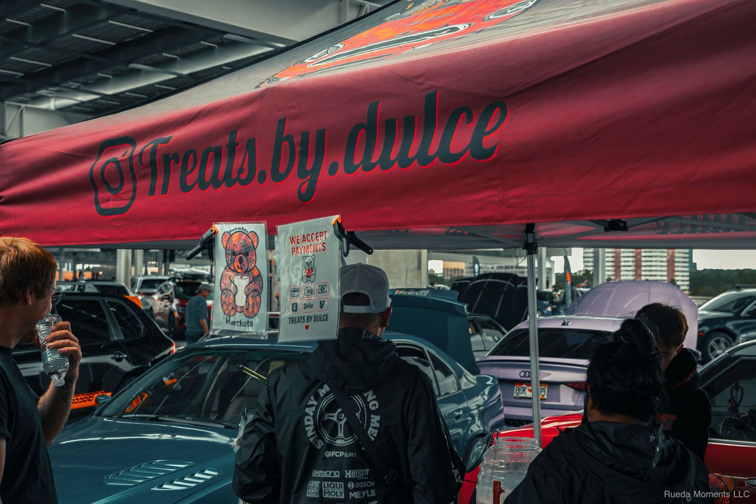 Outdoor car show vendor booth with red @treats.by.dulce banner tent surrounded by modified cars.