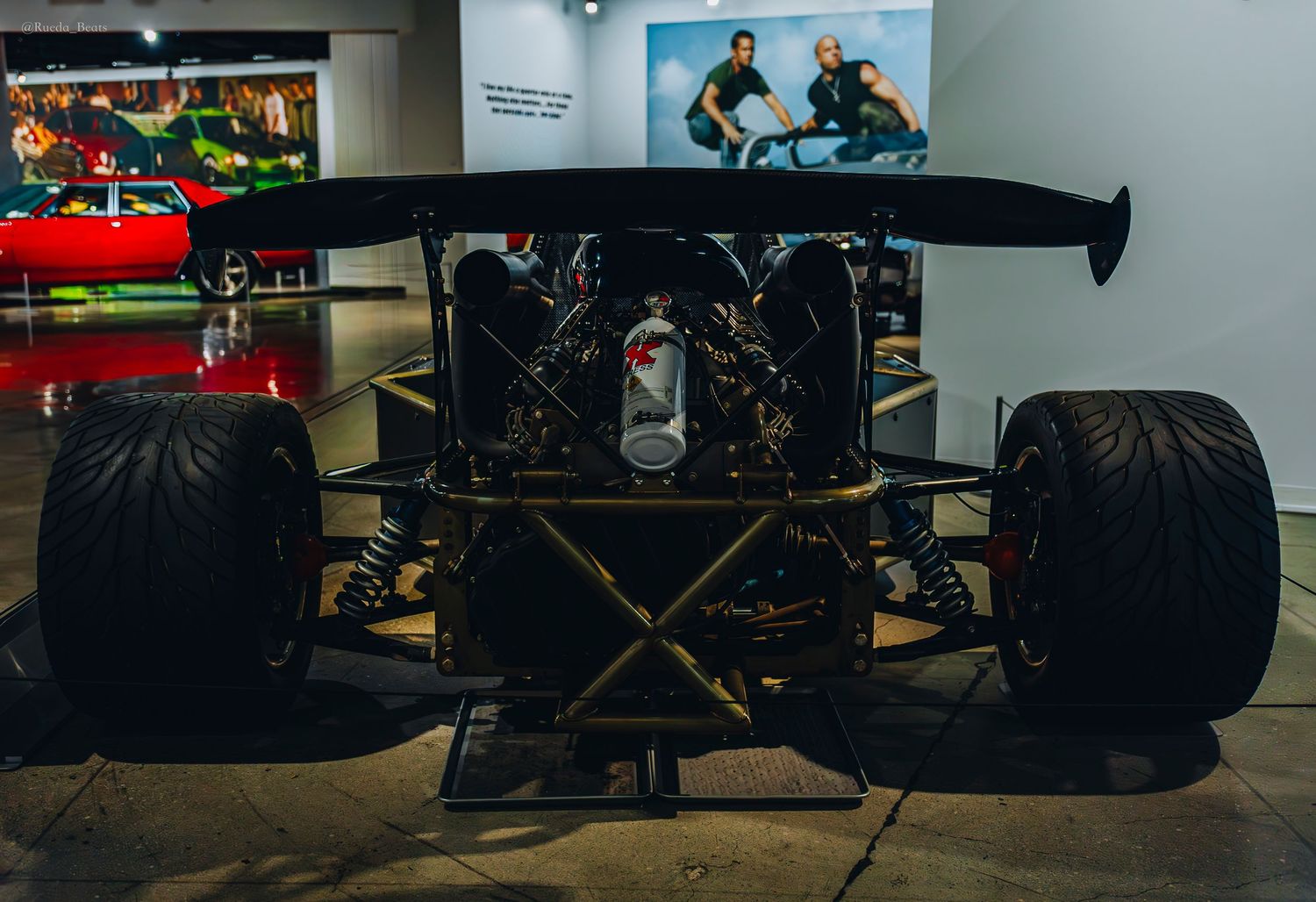 Rear view of black open-wheel race car chassis with large wing and slick tires on display.